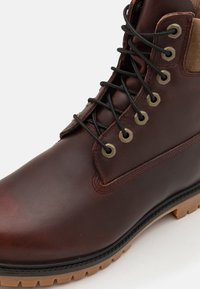 Stivaletto in pelle marrone con lacci neri, occhielli metallici e un accento in suede beige a contrasto. Presenta una suola in gomma robusta con scanalature per la trazione.