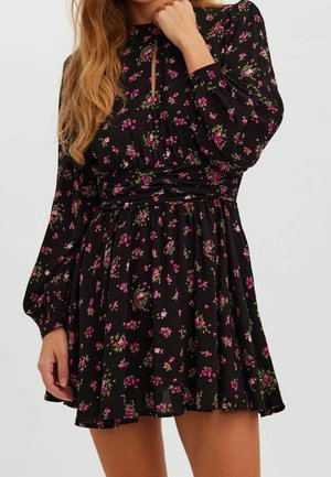 Kobieta w czarnej mini sukience z długim rękawem, ozdobionej różowo-zielonym kwiatowym wzorem, z marszczonymi talią i małym detalem w postaci wycięcia przy dekolcie.