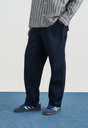 Személy sötétkék nadrágban és kék sportcipőben világoskék csíkokkal, kék szőnyegen áll, kezét a zsebében tartja, részben látható csíkos ing.