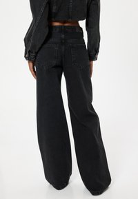 Jeans de mezclilla negra de pierna ancha con una cintura alta, dos bolsillos traseros y una textura sutilmente desgastada. La tela es ligeramente estructurada y gruesa.