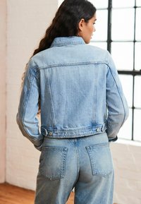 Chaqueta de mezclilla azul claro con un diseño corto, detalles de botones y un cuello clásico. La tela tiene una textura ligeramente desgastada.