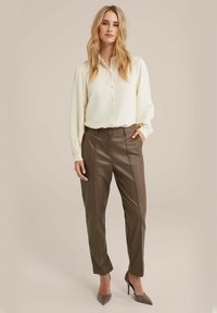 Romantisch knoopshirt in crèmekleur met gestructureerde stof, gecombineerd met hoge bruine faux leren broeken. Neutrale hoge hakken maken de look compleet.