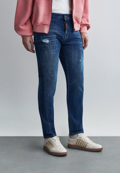 Jean en denim bleu avec détails usés et éclaboussures de peinture, associé à des baskets beige avec des rayures dorées. Sweat à capuche rose clair par-dessus.