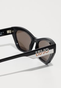 Ochelari de soare negri cu formă de ochi de pisică, cu lentile maronii și brațe din metal argintiu ce poartă logo-ul "Liu Jo" în portocaliu. Material plastic neted, design elegant.