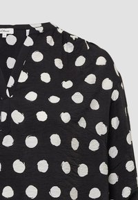 Schwarze Bluse mit V-Ausschnitt und langen Ärmeln, verziert mit unregelmäßigen weißen Punkten in verschiedenen Größen, aus leichtem Stoff gefertigt.