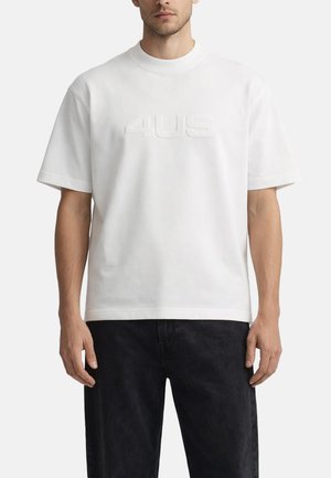 Uomo che indossa una t-shirt bianca semplice a maniche corte con il logo "4US" in rilievo sul petto, abbinata a pantaloni neri su sfondo bianco.