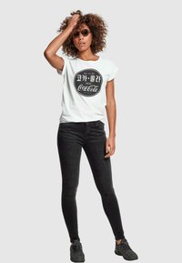 Un tricou alb cu mânecă scurtă, cu un logo Coca-Cola rotund în text negru, asociat cu blugi negri slim fit și adidași negri texturați.