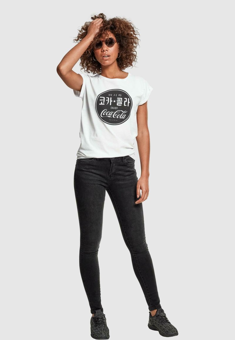 Un tricou alb cu mânecă scurtă, cu un logo Coca-Cola rotund în text negru, asociat cu blugi negri slim fit și adidași negri texturați.