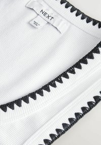 Vit ribbad tygblus med svart zigzag-stickning längs kragen. Etikett läser "NEXT", tillverkad i Indien. Texturerad yta.