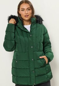 Grön puffjacka med fake päls på huven, med dragkedja och tryckknappar, kviltat mönster och två sidofickor. Slät, glänsande yta.