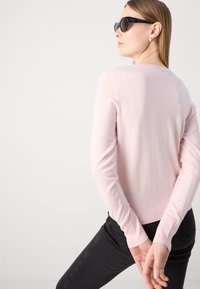 Vaaleanpunainen neulepusero pitkillä hihoilla ja ribbaamalla viimeistellyllä helmalla, yhdistetty mustiin farkkuihin. Sujuva pinta, istuva muotoilu ja pyöreä pääntie.