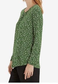 Femme portant un chemisier vert à manches longues avec de petits motifs floraux blancs, associée à un pantalon noir, sur un fond uni.