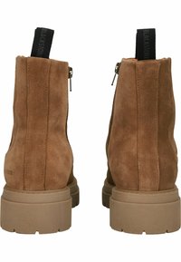Botas de ante marrón hasta el tobillo con una textura suave, cremalleras laterales y una suela de goma texturizada. Presenta una lengüeta para tirar en bucle con una marca negra.