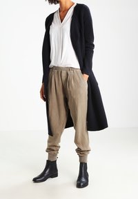 Larga chaqueta de punto color marino, camisa blanca de cuello en V, pantalones beige relajados y botas negras hasta el tobillo. Enfoque en la tela en capas y la paleta de colores neutros.