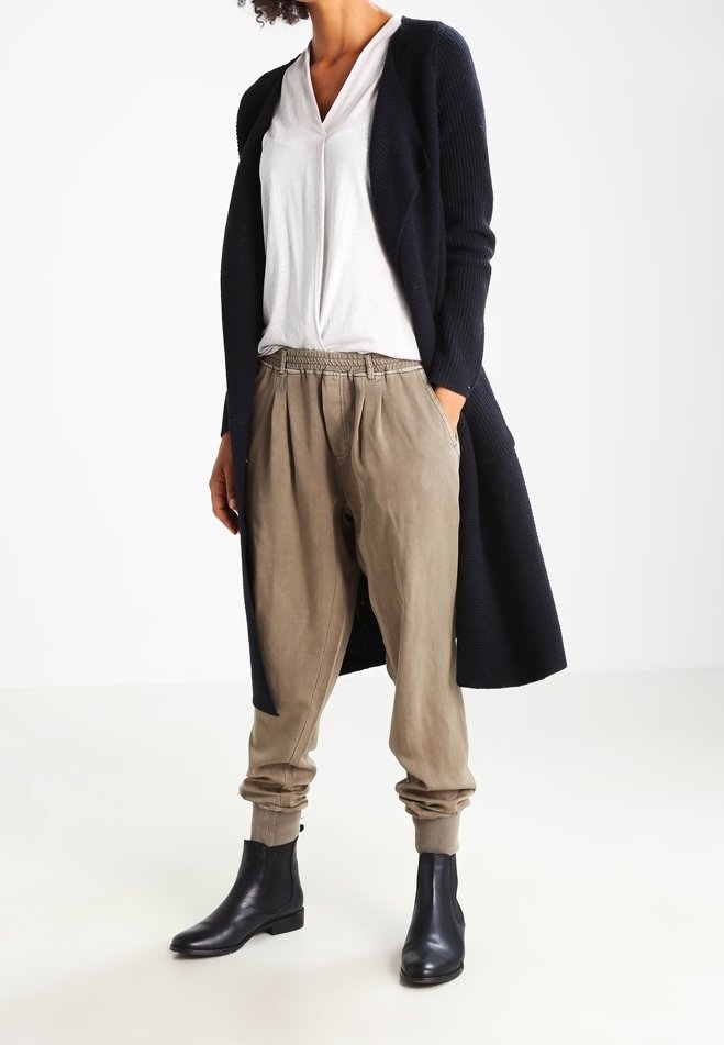 Larga chaqueta de punto color marino, camisa blanca de cuello en V, pantalones beige relajados y botas negras hasta el tobillo. Enfoque en la tela en capas y la paleta de colores neutros.