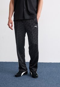 Sorte sweatpants med et gentagende logo, prydet med hvide side-striber og lavet af glat materiale; båret med sorte løbesko.