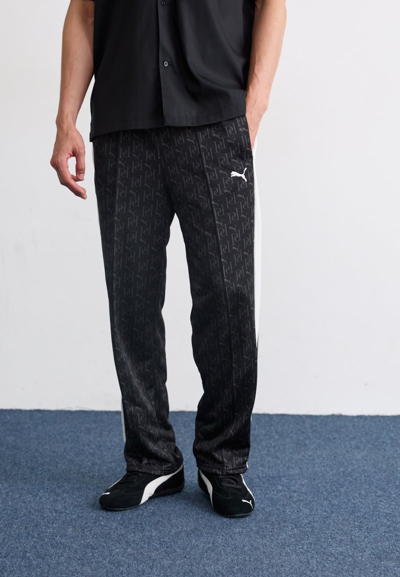 Sorte sweatpants med et gentagende logo, prydet med hvide side-striber og lavet af glat materiale; båret med sorte løbesko.
