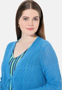 Hellblauer Strickcardigan mit offenen Mustern und einer Bindeschleife an der Vorderseite, über einem blau-grün gestreiften Oberteil getragen.