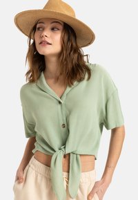 Camisa de botones de manga corta en verde menta atada a la cintura, hecha de tejido texturizado, combinada con pantalones beige claros y un sombrero de paja.