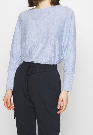 Personne portant une chemise à manches longues bleu clair rentrée dans un pantalon noir à cordon avec des poches cargo, debout devant un fond uni.