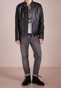 Schwarze Lederjacke mit hohem Kragen, Reißverschluss und zwei vorderen Taschen, kombiniert mit einem grauen T-Shirt und grauen Jeans, die über dem Knöchel enden.