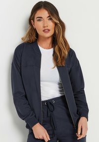 Marineblauwe bomberjack met rits, voorzien van ribbelmanchetten en -kraag, gemaakt van zachte stof. Gecombineerd met een wit T-shirt en bijpassende marineblauwe broek.