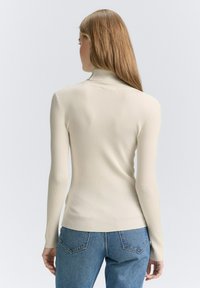 Tricou cu guler înalt și mâneci lungi, cu textură ribbed, de culoare bej deschis, având un design slim și o textură netedă, asortat cu blugi albastri cu talie înaltă.
