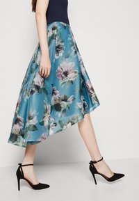 En medellång blommig kjol i turkos och vit, med en plisserad design och parat med svarta högklackade skor. Slät textur.