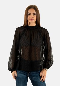 Blusa negra transparente con estampado de lunares, cuello con volantes, mangas abullonadas y corte holgado, combinada con jeans de mezclilla oscuros.