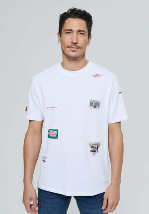 T-shirt en coton blanc à manches courtes, ornée de plusieurs patchs colorés et de textes. Coupe régulière avec un col rond et une texture douce.