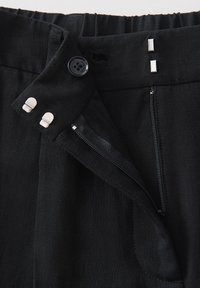Black trousers with a textured fabric, featuring a button, zipper closure, and two silver accent pieces at the waistband.