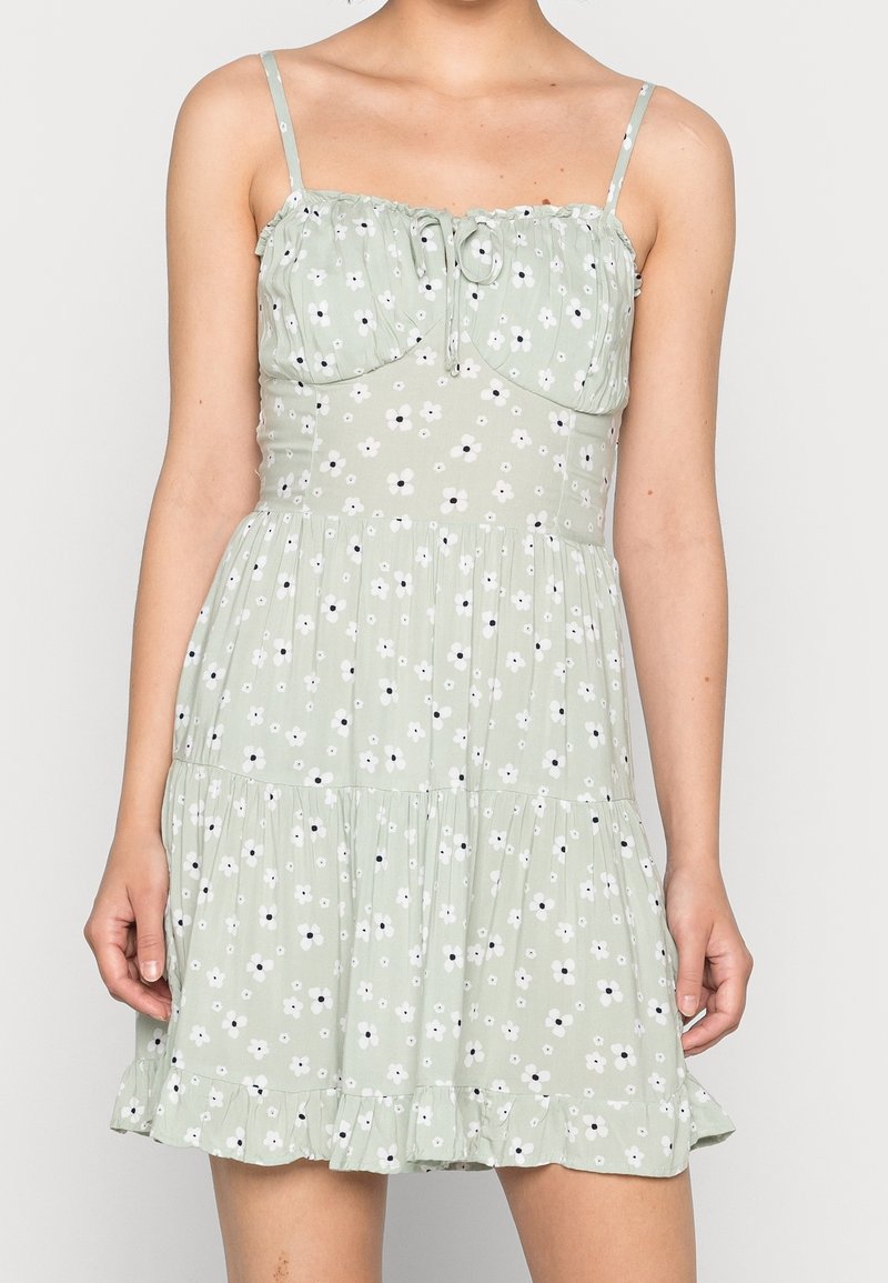 Grön blommig klänning med tunna band, volangkant och skräddad byst som har vita blommor med svarta mittpunkter. Tillverkad av lätt tyg.