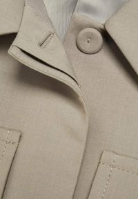 Gros plan d'une veste en tissu beige montrant un bouton, une boutonnière, le bord du col et les détails de couture de la poche.