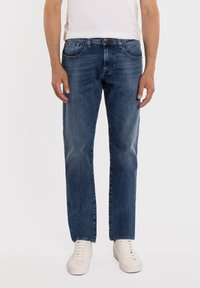 Mężczyzna ubrany w wyblakłe, niebieskie jeansy o prostych nogawkach, białą koszulkę i białe trampki, stojący z ramionami swobodnie opuszczonymi wzdłuż ciała.