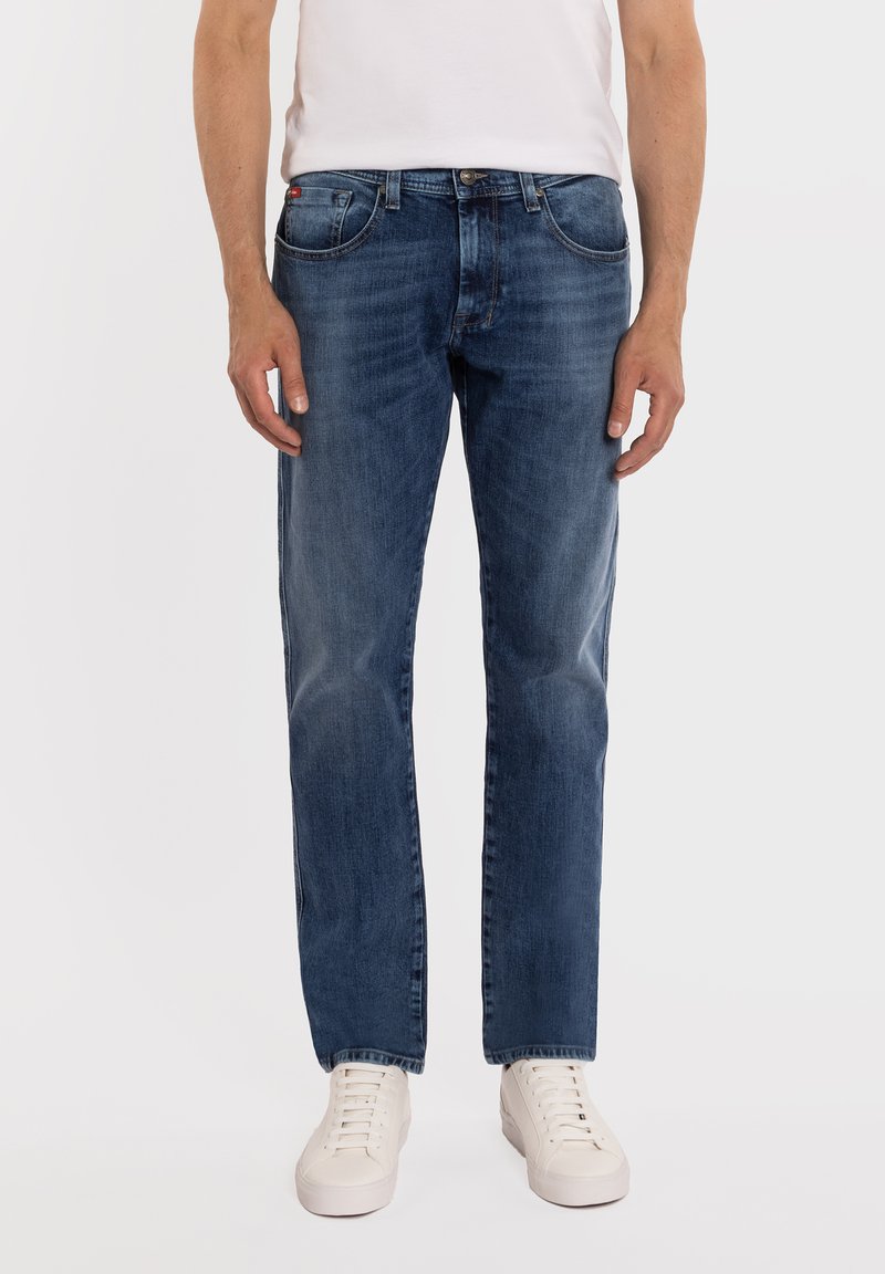 Mężczyzna ubrany w wyblakłe, niebieskie jeansy o prostych nogawkach, białą koszulkę i białe trampki, stojący z ramionami swobodnie opuszczonymi wzdłuż ciała.