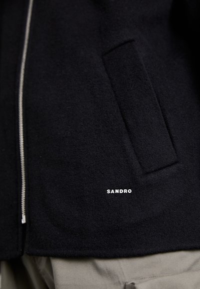 Juodas vilnonis džemperis su sidabriniu užtrauktuku, turintis šoninį kišenę ir baltą "SANDRO" logotipą priekyje. Lygus paviršius.