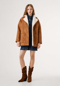 Cappotto in suede marrone con colletto in pelliccia sintetica bianca, caratterizzato da una vestibilità ampia e chiusura con bottoni, indossato sopra un vestito in denim e stivali in suede marrone.