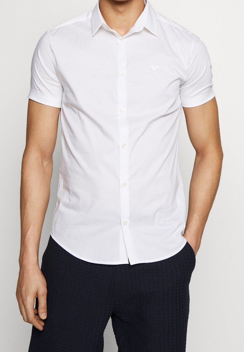 Vit kortärmad skjorta med knappar, tillverkad av mjukt tyg, med en standardkrage och en diskret logotyp på bröstet.