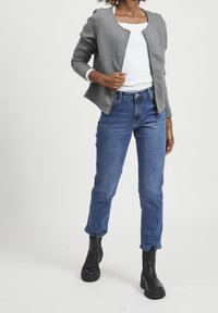 Šedá texturovaná bunda s kulatým výstřihem, v kombinaci s bílým topem a modrými rovnými džíny. Černé kotníkové boty dokončují outfit.