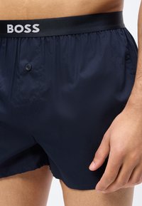 Homme portant un boxer bleu marine foncé à braguette boutonnée et ceinture noire portant l'inscription "BOSS", montrant le torse et la main droite.