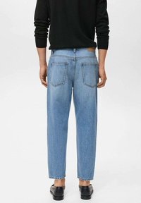 Una persona è in piedi di spalle, indossando un maglione nero e jeans azzurri chiari, abbinati a scarpe nere. Lo sfondo è semplice e chiaro.