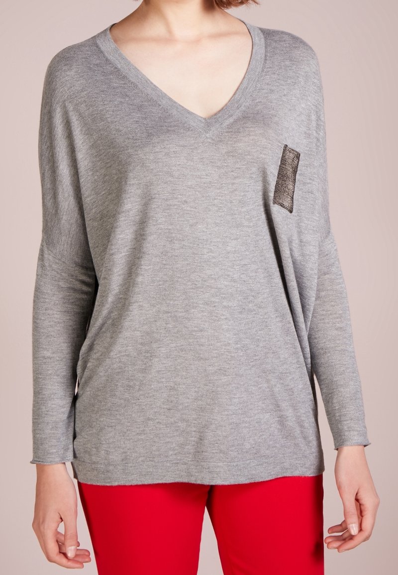 Grauer V-Ausschnitt-Pullover mit lockerer Passform, langen Ärmeln und einer strukturierten schwarzen Tasche. Weicher Stoff, legeres Design, kombiniert mit roter Hose.