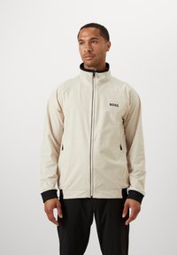 Hellbeige Zip-Jacke mit hohem Kragen, schwarzen Akzenten, zwei seitlichen Taschen und dem Markenzeichen "BOSS" auf der Brust. Glatte Textur.