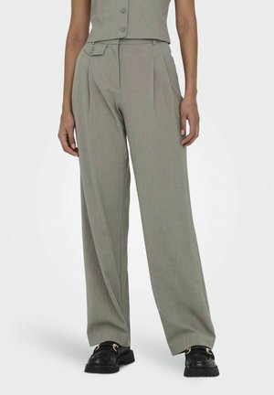 Femme portant un pantalon gris taille haute à jambes larges avec une patte de taille boutonnée et des mocassins noirs, debout devant un fond uni.
