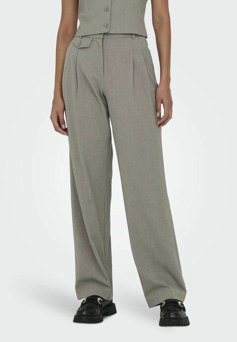 Femme portant un pantalon gris taille haute à jambes larges avec une patte de taille boutonnée et des mocassins noirs, debout devant un fond uni.