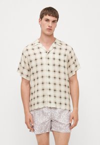 Camicia a maniche corte, con bottoni, in un motivo a quadri beige e nero, realizzata in tessuto leggero con una vestibilità rilassata. Il modello indossa pantaloni corti grigi con motivo logo.