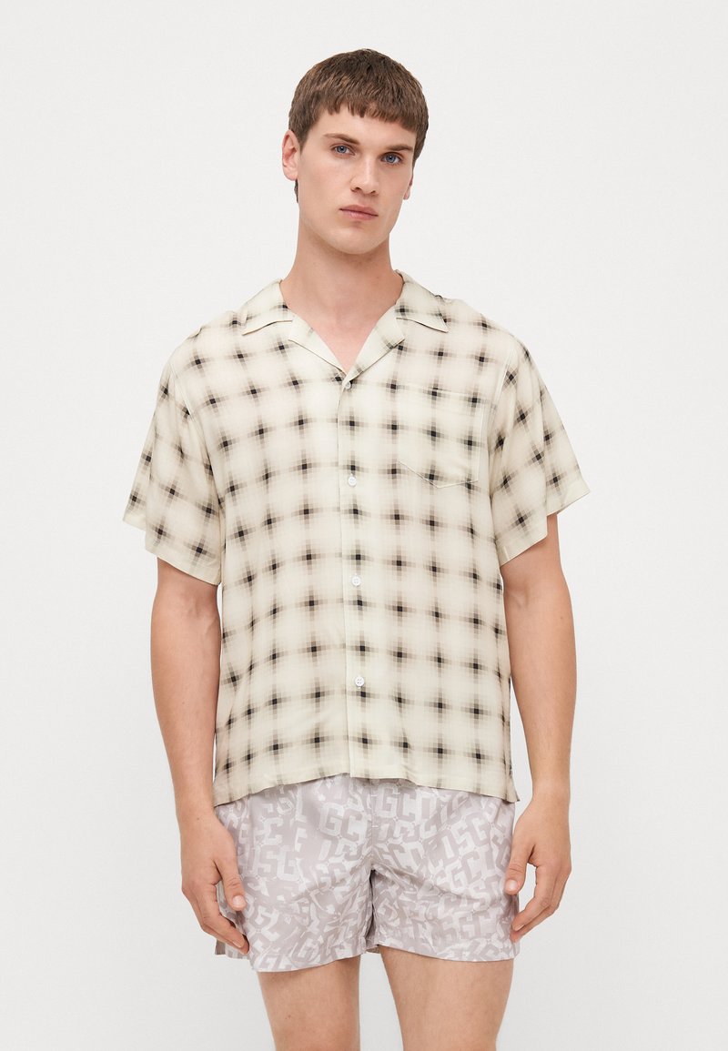 Camicia a maniche corte, con bottoni, in un motivo a quadri beige e nero, realizzata in tessuto leggero con una vestibilità rilassata. Il modello indossa pantaloni corti grigi con motivo logo.