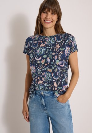Lachende vrouw met bruin haar die een marineblauw shirt met korte mouwen draagt met meerkleurige bloemmotieven en een lichtblauwe spijkerbroek met hand in de zak.