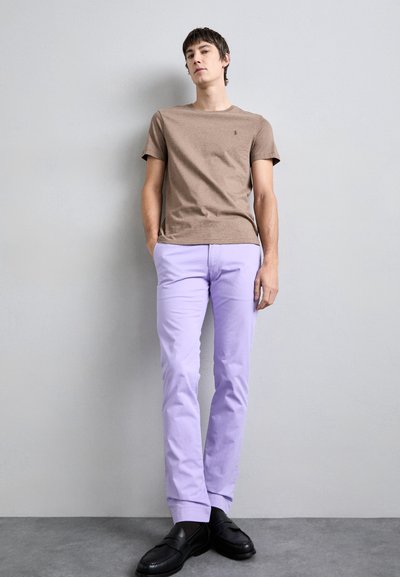 Camiseta de algodão castanha, com mangas curtas, combinada com calças de algodão lilás claras e mocassins pretos. O modelo está de pé contra um fundo cinza.