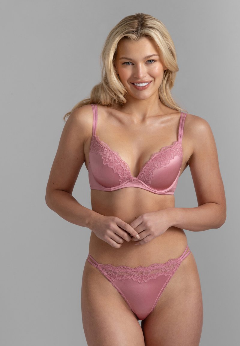 Γυναίκα με μακριά ξανθά μαλλιά που χαμογελά, φορώντας ροζ σουτιέν με δαντέλα και ταίριαστα εσώρουχα, μπροστά σε απλό γκρι φόντο.